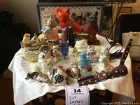 Overview of entire cat lovers lot showing all figurines, music boxes, salt and pepper shakers, pincushion, print, and book on a table with white cloth.