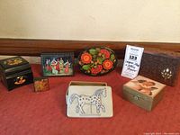 Full view of seven decorative boxes including hand-painted lacquer, wood inlay, paper mache, and pottery box arranged on red cloth with measurement scale visible