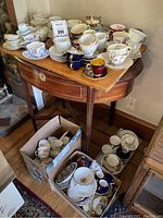 Wide view showing numerous tea cups and saucers arranged on a wooden table with some items also in cardboard boxes on the floor. The collection is assorted in style and design.
