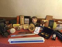 Wide overview showing vintage Kodak Jr camera, multiple rulers and scales, political pin badges, letter openers, paperweights, and novelty items arranged on a cloth.