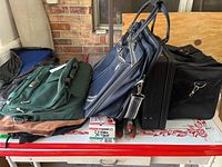 Assorted travel and camera bags including navy blue Eddie Bauer soft-side rolling luggage, black hard-side briefcase with combination lock, green fabric bag, and black camera case with handles, placed on table.