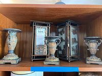 Wood shelf displaying three urn-shaped candle holders flanking two rectangular lanterns with butterfly and floral designs on glass panels.
