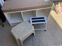 Front view of white mud-room cubby bench with tan cushion and two step stools: one plastic, one metal frame.