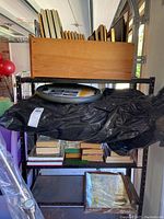 Various picture or plaque frames with protective glass arranged upright in a wooden box on a black metal shelf with other items beneath.
