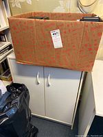 White MDF storage cabinet with two doors and two handles, placed on floor with a cardboard box and trash bag nearby