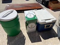 Three items arranged on pavement: green Igloo water cooler, boxed patio set cover, blue and white Coleman chest cooler