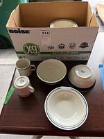 Six stoneware and ceramic dishes including coffee cups and bowls in cream color, some upside down, inside and next to a cardboard box.