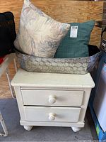 White painted two-drawer nightstand with round knobs, scratches visible on surface; large aluminum decorator tub with wire mesh atop the nightstand filled with two different throw pillows, one floral pastel and one solid blue-green.