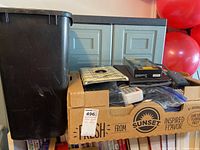 Photo showing a cardboard box filled with phone cases, phones, calculator, a black plastic trash can beside the box, and VHS tapes below the box