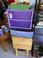 Photo showing two sets of plastic storage bins/drawers stacked, one purple with rollers and one with black frame and translucent drawers, with two wooden fold-out TV trays below them.