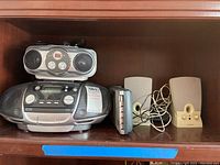 Photo showing several audio devices: RCA boombox with radio, CD and cassette capabilities, Insignia CD player, Radio Shack cassette recorder/player, and the pair of Hartman/Kardon computer speakers, beige color with wires.