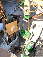 Scott’s Snap lawn feeder with green and beige chute and wheels, along with a box holding garden tools and trimmer attachments placed on ground outdoors.