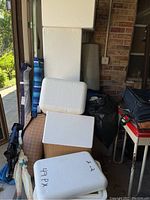 Stack of white styrofoam coolers and lids, some labeled with markers, placed beside various bags and boxes.
