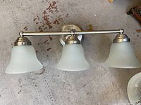 Three-light brushed nickel finish wall light fixture with frosted bell-shaped glass shades resting on floor.