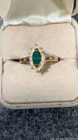 Front view of gold-tone ring with green marquise center stone surrounded by clear stones set on a size 4 band inside beige fabric-lined box.