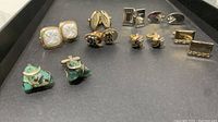 Eight pairs of vintage cuff links displayed on dark background showing variety in styles and materials including mother-of-pearl, green marbled stone, gold-tone metal, and textured patterns.
