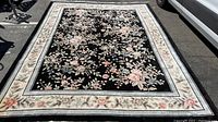 Full view of large rectangular area rug with black center and floral ivory border laid on pavement.