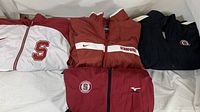 Photo of four folded windbreaker jackets in various Stanford-themed designs and colors, including red, white, and navy blue.