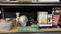 Wide view of the shelf showing boxed Braun juicer and Hamilton Beach hand blender, Cephalon serving tray, ceramic chef figurine, glasses, aluminum mold, and decorative glass bowl.