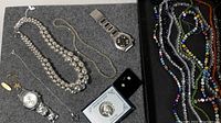 Wide view showing two men's wristwatches, two large silver-tone bead necklaces, a thin gold chain necklace, a delicate pendant necklace, small earrings, and a silver commemorative half-dollar coin in a protective case.
