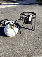 Photo showing black metal outdoor stove with three legs and a circular ring on top, connected via hose to a white propane tank on pavement.