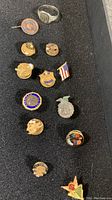Photo showing collection of 12 vintage service pins and one vintage ring on black background. Pins include police badge, American flag, eagle emblem, and several gold-tone and enamel pins with various insignias and colors including blue, red, and green. Ring is silver-tone signet style, dated wear seen on all items.