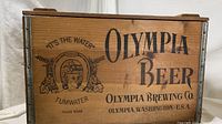 Side view of wooden Olympia Beer crate showing branding and graphic of horseshoe with hops and barley and 'It's The Water' slogan.