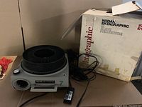 Photo of the Kodak Ektagraphic III slide projector with carousel on top, wired remote beside it, power cord, and the original box in the background.