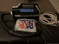 Oreck XL handheld vacuum cleaner alongside a long white power cord and a pack of Oreck vacuum bags on a dark surface.