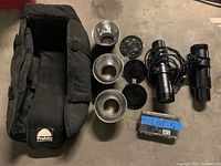 Lot overview showing two Profoto Compact lights, several metal reflectors, honeycomb grids, a carrying bag, and a small accessory case on the floor.