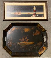 Photo showing framed sailing ship print featuring lighthouse on the right and sailing ships on the left under a starry sky, above a vintage metal serving tray decorated with gold-tone sailing ship and floral stencils on a dark background.