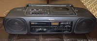 Front angled view of black Sony boom box showing speakers, cassette deck, buttons, and top-loading CD player lid.