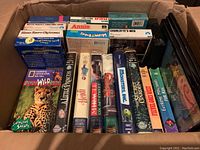 Photo of a cardboard box filled with various children's VHS tapes and a few blank VHS cases visible on the right side. Titles include Elmo Saves Christmas, Annie, Charlotte's Web, A Little Princess, Beasties of the Wild, Monsters Inc., Ernest Scared Stupid, Home Alone 2, National Geographic Dancing Safari among others.