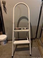 Front view of white metal step stool showing two wide steps, curved handle, and safety instruction label on top step.
