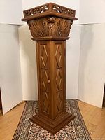 Front view of the carved wooden pedestal showing detailed carvings, diamond pattern door, and shaped base.