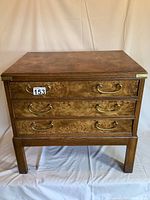 Front view of the campaign style wooden chest with three drawers and brass handles sitting on a wooden base.