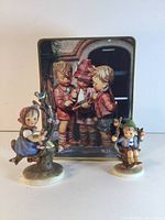 Photo showing the two Hummel apple tree-themed figurines and the metal box with Hummel artwork on the lid arranged together.