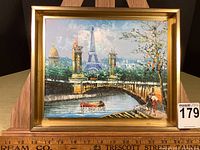 Frontal view of the painting showing the Eiffel Tower, a bridge, a boat in the river, and two people walking along the riverbank under trees. The painting is framed in a gold-colored frame. A ruler at the base shows the size approximates 10x12 inches including frame.