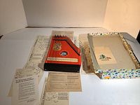 Overview photo showing the red Jubel Tone zither with 27 strings placed on a table surrounded by multiple sheets of instructional music paper and the original colorful box.