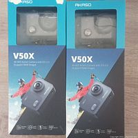 Front view of two factory sealed Akaso V50X 4K Wi-Fi action camera boxes showing the camera visible in plastic display window and packaging design with skier and camera features.