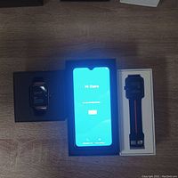 Three boxes shown on a wooden surface. The smartphone is shown powered on in center box, flanked by two smartwatches each in their respective boxes.