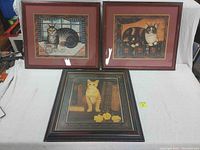 Three framed cat prints displayed together on white fabric background, two with red wood frames and one with dark wood frame.