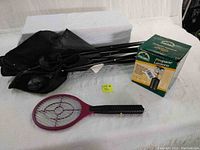 Photo showing collapsed black camping chair, electric fly swatter (purple racket) and boxed propane lantern