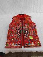 Front facing image of a red sleeveless vest with collar on a hanger laid on a white background. The vest is richly embroidered with colorful circular and floral patterns. Zipper and collar are visible.
