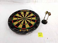 Winmau official British Darts Organization dartboard displayed flat, showing segment colors and numbering. Two darts with large flights are held upright in a small round black stand next to it.