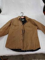 Full front view of the brown men's work jacket laid flat, showing two chest flap pockets, button front, collar, and signs of wear including small stains.