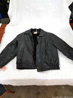 Front view of the black leather jacket laid on a white background, showing full length, collar, pockets, and zip-up front.