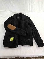 Black men's cable knit sweater spread flat showing front, button-down style, textured knit, brown elbow patches, and shawl collar.