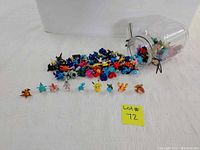 Photo showing a large glass jar tipped over with many small Pokemon figurines spilling out. Several figurines are lined up in front, including Pikachu, Charizard, and other Pokemon characters in various colors.