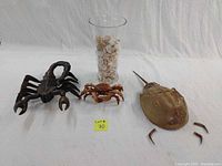 Photo showing all four items: large horseshoe crab with detached legs, wooden crab figurine, wooden scorpion, and glass vase filled with shells on white background.
